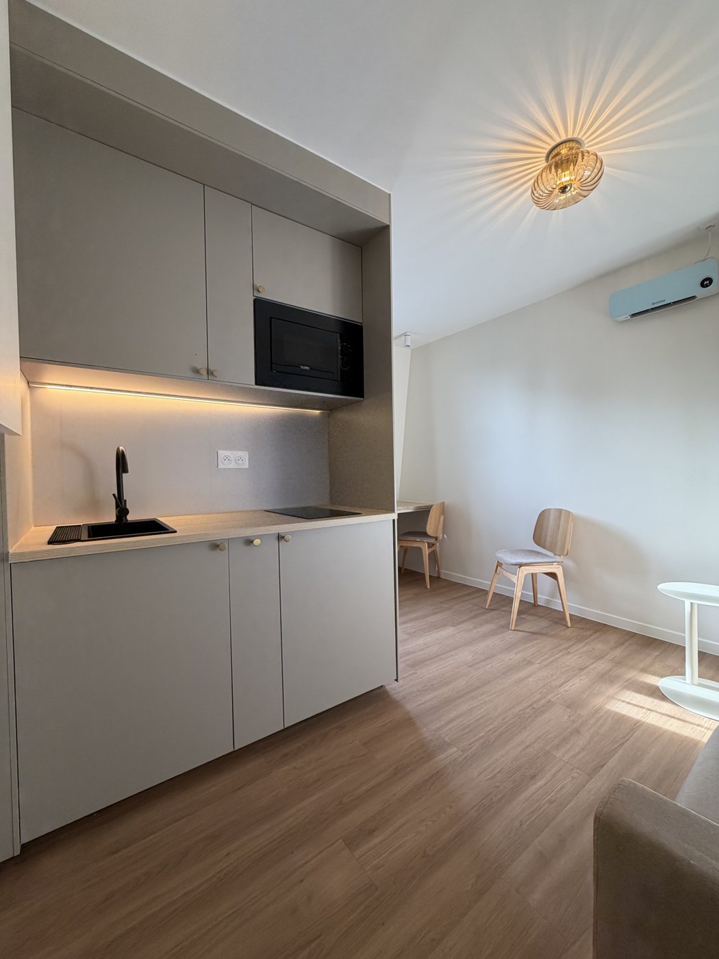 Cuisine intégrée du studio aux façades grège, sous un luminaire doré et une lumière chaude